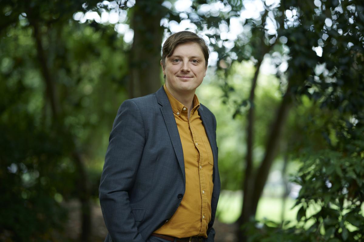 Image of Nick Keegan in a yellow shirt and grey suit, with trees in the background