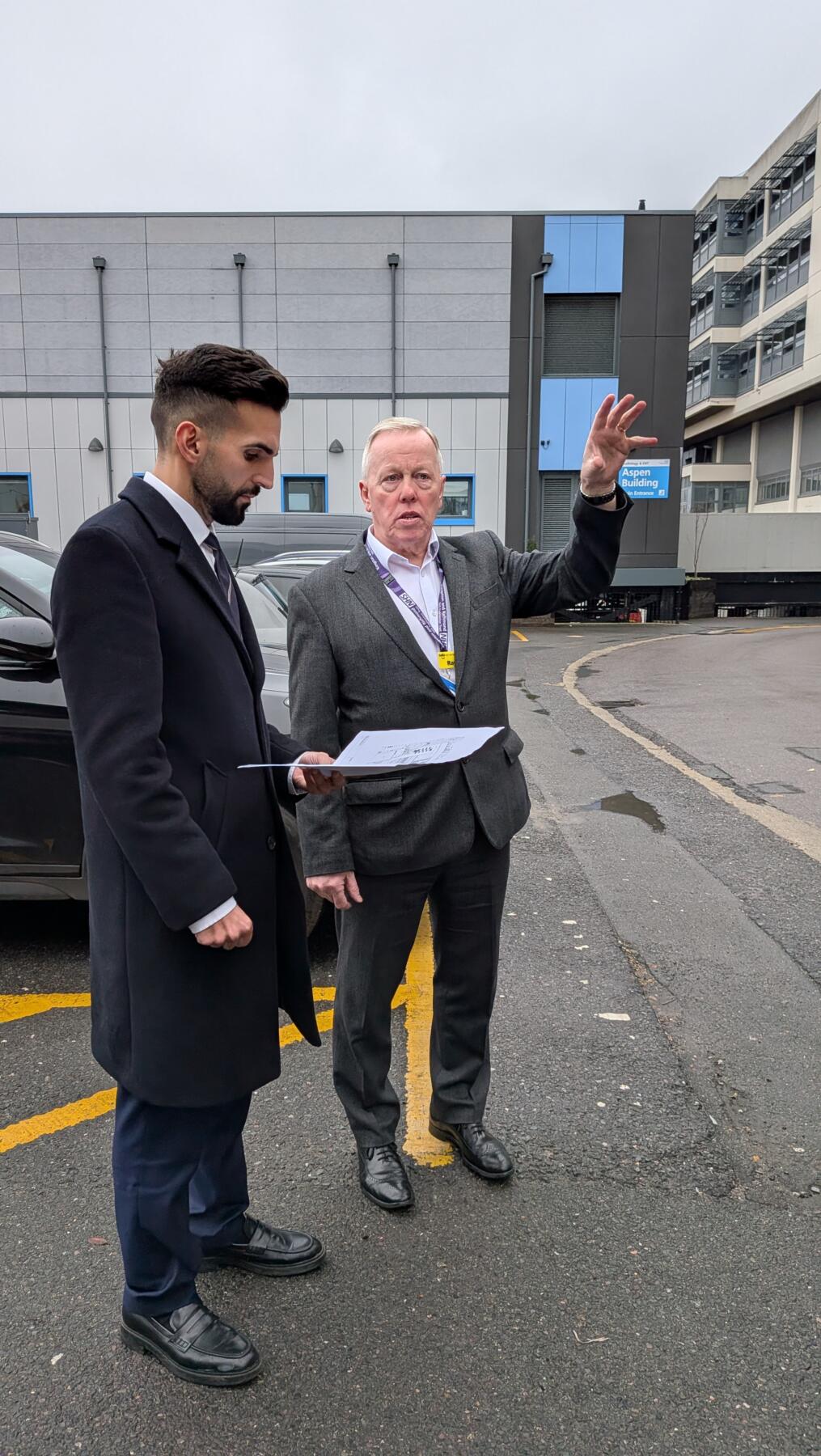 Two people standing in a car park; one is looking at a printed building plan and the other is gesturing