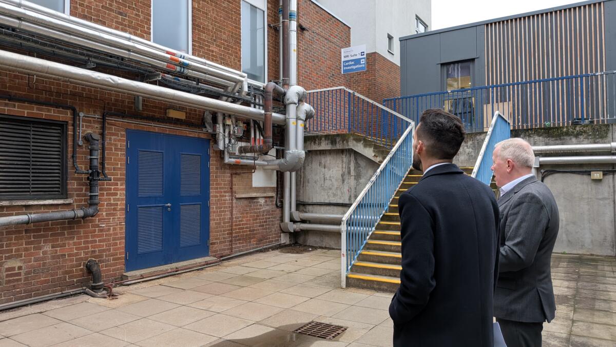 Two men looking at a building with insulated heating pipes running along a brick wall near a blue plant‑room door and a staircase leading to an upper level. 