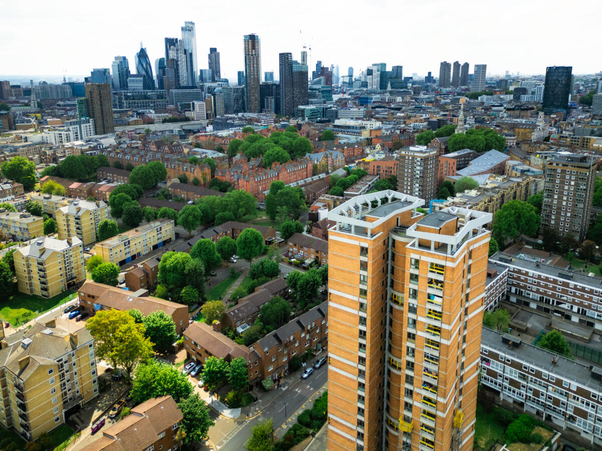 Aerial view of a urban neighbourhood with mid-rise residential buildings and tree-lined streets in the foreground, leading to the City of London skyline in the background, including distinctive skyscrapers such as the Gherkin, under a bright overcast sky