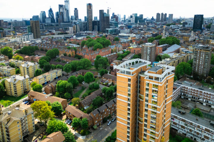 Aerial view of a urban neighbourhood with mid-rise residential buildings and tree-lined streets in the foreground, leading to the City of London skyline in the background, including distinctive skyscrapers such as the Gherkin, under a bright overcast sky