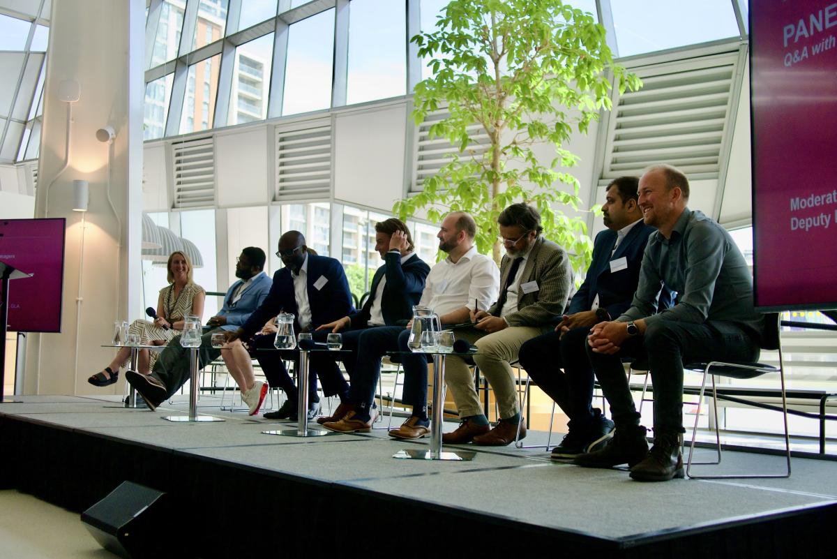 Panel discussion on a stage in a glass-walled venue, with eight seated speakers, small tables, water jugs and display screens.