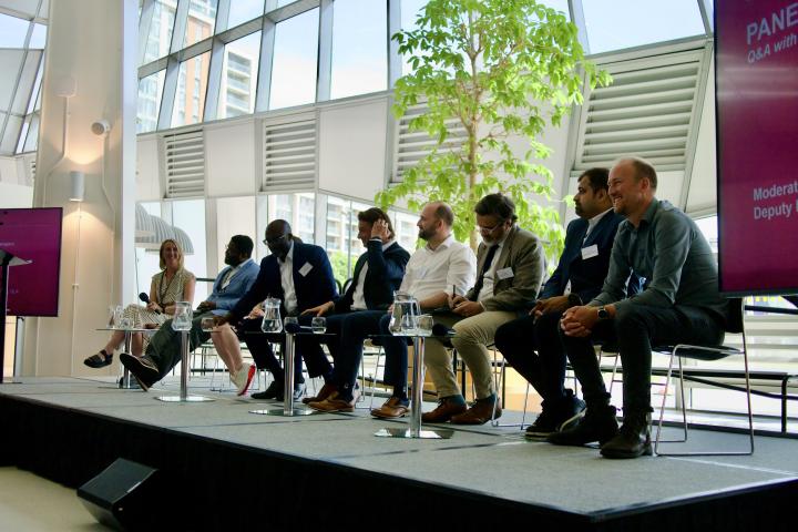 Panel discussion on a stage in a glass-walled venue, with eight seated speakers, small tables, water jugs and display screens.
