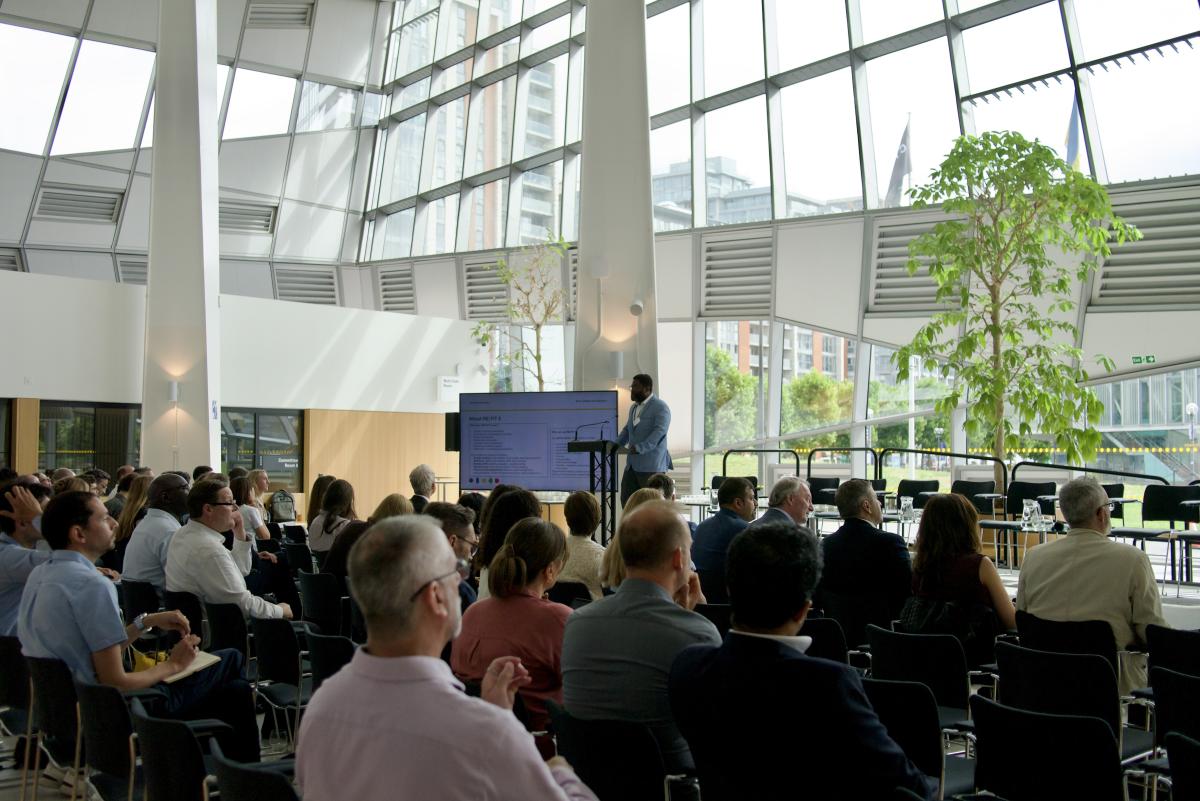 Audience seated in a bright glass-walled venue watching a speaker at a lectern beside a presentation screen.