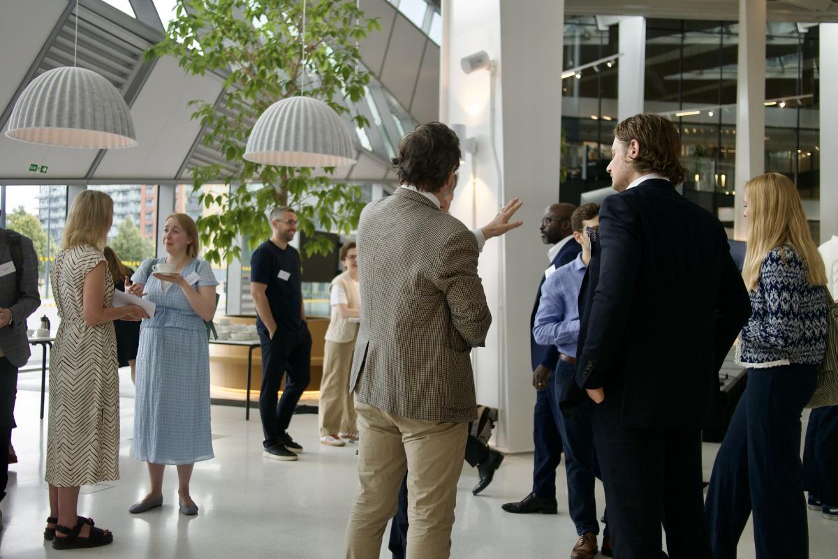 Attendees stand and talk in a bright atrium with large windows, indoor trees and hanging lights during an event networking session.